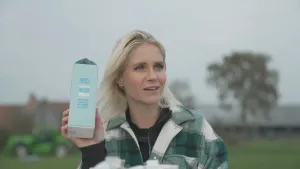 Een foto van Janouk op een grasveld. Ze houdt een pak Kokosmelk vast in haar rechterhand en kijkt naar rechts naar de niet-zichtbare Casper. Op het pak staat dat het drinken 100% plantaardig is, lactose en glutenvrij en calcium, Vitamine B12 en D2 bevat.
