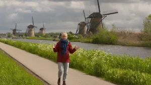 Een foto van Janouk die over een pad langs het water loopt en haar handen uit elkaar houdt voor haar lichaam. Aan de andere kant van het water staan vijf molens met ieder vier wieken. Het zijn de molens van Kinderdijk, die beschermd werelderfgoed zijn.