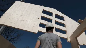 Een foto van een houten zijkant van een woning. Dat paneel wordt opgetild door een bouwkraan. Op de voorgrond zie je een bouwvakker met een helm op. 