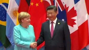Een foto van de voormalige leider van Duitsland, Angela Merkel en de leider van China Xi Jinping. Ze schudden elkaar de hand. Op de achtergrond zie je allemaal vlaggen van landen. 