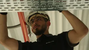 Een foto van een man die aan het werk is. Hij heeft een bouwhelm op. Je ziet een ijzeren plaat en hij doet iets op die plaat, maar dat is buiten beeld.