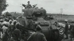 Een foto van een tank in Nederlands-Indië. Het is een zwartwit-foto. De tank rijdt op een weg en langs de weg staan allemaal mensen. Erachter rijdt nog een tank. 