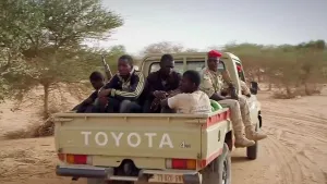 Een foto van een savanne in Afrika. Je ziet een rijdende pick-up truck met in de laadbak vijf mannen. Twee van die mannen hebben een geweer. En een van hen heeft een uniform aan.