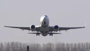 Een foto van een vliegtuig dat aan het opstijgen is. Het vliegt in de richting van de camera. Onderin beeld zie je een hek en in de verte zie je een rij bomen.