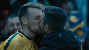 Een foto van twee mannen die elkaar op de mond zoenen. Van een man is het duidelijk dat hij een voetbalshirt aan heeft. Op de achtergrond zie je nog een man.