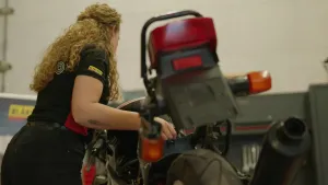 Een foto van een vrouw die werkt aan een motorfiets. Ze heeft zwarte kleren aan en staat in een werkplaats.