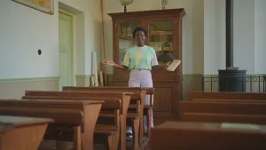 Een foto van presentator Tirsa. Ze staat in een heel oud klaslokaal en heeft haar armen gespreid. Ze heeft een boek vast en kijkt in de camera. 