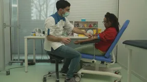 Een foto van presentator Eva. Ze zit in een stoel bij de dokter. De dokter zit tegenover haar en drukt met zijn wijsvinger op de onderarm van Eva. De dokter draagt een mondmasker en heeft een naald in zijn andere hand.