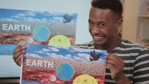 Een foto van een presentator die lacht. Op de papieren vlag die hij vastheeft staat de tekst "Earth", foto's van de vier elementen lucht, aarde, vuur en water en tekeningen van de wereldbol, een vredesduif met olijftakje en een man, de Vitruviusman.