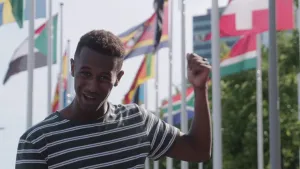 Een foto van een presentator die buiten met zijn linkerduim naar achteren wijst. Achter de presentator staan acht vlaggen van landen op een witte vlaggenstok. Eén vlag is goed te zien en die is van Zwitserland: een wit plusteken op een rode achtergrond.