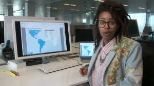 Een foto van presentator Eva in een jasje met de wereldkaart erop. Op het bureau achter haar staat een beeldscherm en een laptop waarop een blauwe kaart staat met daarop alle ambassadeurs van Nederland in de Verenigde Staten van Amerika en Zuid-Amerika. 