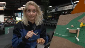 Een foto van Janouk in een zwart en blauw pak naast een lichtbruin bord met gaatjes. Het bord kan tonen hoe een seintje van pijn doorgegeven wordt. Op een groen getekende hand kan iets vallen in een buisje, vallen op een bakje en een aan-uit knop raken.