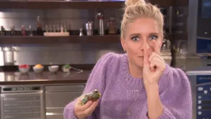 Een foto van Janouk in een keuken. Ze heeft in haar rechterhand een rolletje met bij elkaar gerolde eetbare microgroenten vast. Ze houdt haar linkerduim en linkerwijsvinger iets uit elkaar voor haar gezicht, om aan te geven hoe klein microgroente is.