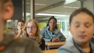 Een foto van een schoolklas waarin vier kinderen deels te zien zijn en presentator Sosha rechtsachterin zit. Ze luisteren met aandacht naar de gastleraar Lucelle Comvalius voor de klas buiten beeld. Lucelle is aan het vertellen over racisme.