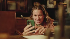 Een foto van presentator Pascal die achter een bureau zit en zijn mobiele telefoon in de hand heeft. Hij leest van het scherm informatie op over wat een QR-code is. Er is geen QR-code zichtbaar en Pascal zal er later in de uitzending één gaan scannen.