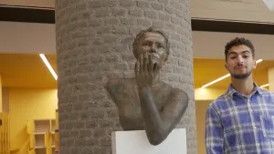 Een foto van presentator Nizar. Hij staat rechts in beeld en kijkt in de camera. In het midden zie je een standbeeld van Marga Klompé. Daarachter zie je een brede pilaar. In de achtergrond zie je kluisjes.