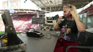Een foto van presentator Janouk. Ze zit in een voetbalstadion, tussen de tribunes, achter een tafel. Ze heeft een headset op. Op de achtergrond zie je een Tv-camera. Ze kijkt in de richting van het veld. 