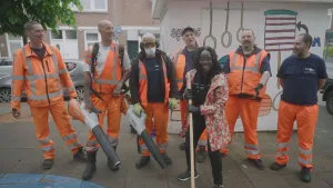 Een foto van presentator Tirsa die op straat staat met een bezem in haar handen. Achter haar staan zes professionele afvalophalers. Ze hebben opvallende oranje kleren aan. Twee van hen hebben een bladblazer vast.