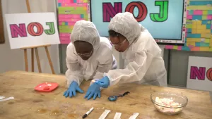 Een foto van Tirsa en Eva die een dun wit veiligheidspak dragen, plastic blauwe handschoenen en een doorzichtige veiligheidsbril. Ze staan gebogen over een tafel met kleine plastic bakjes met vloeistof, een stopwatch en een schaal met spekjes erin. 