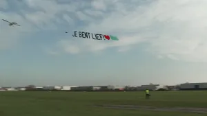 Een foto van een vliegtuig in de lucht met een doorzichtige strook met tekst erachter. Er staat op "Je Bent Lief!" en erachter staat een rood hartje en een groene rechthoek. Er worden ook wel reclames aan een vliegtuig gehangen, dat heet luchtreclame. 