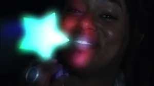 Een foto van het gezicht van presentator Eva die in het donker een groen oplichtende glow in the dark ster aan een puntje van de ster vastheeft. Eva lacht en kijkt naar de camera. De glow in the dark ster bevat stofjes die licht opnemen en afgeven.