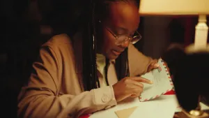 Een foto van presentator Tirsa die aan een tafel met enveloppen erop zit en een envelop waar een brief in kan vastheeft. Ze wijst op het adres dat met pen op de envelop is geschreven. Het adres is niet leesbaar op de foto. 