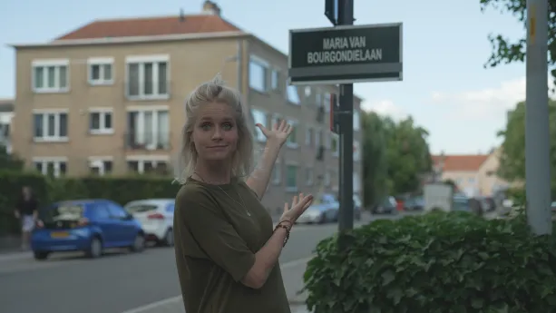 Een foto van presentator Janouk. Ze staat buiten op straat en wijst naar een straatnaambord. Daarop staat "Maria van Bourgondiëlaan". 
