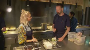 Een foto van presentator Janouk in een professionele keuken. Ze is in gesprek met sterrenkok Emile van der Staak. Op de voorgrond zie je bakjes en flesjes met etenswaren. Op de achtergrond zie je nog twee andere koks staan.