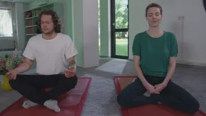 Een foto van presentator Pascal. Hij zit in kleermakerszit op een matje in een woning. Hij heeft geen schoenen aan. Naast hem zit een vrouw, in ongeveer dezelfde houding, ook op een matje. Beiden hebben hun ogen dicht. Ze zitten er heel ontspannen bij.