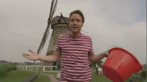 Een foto van presentator Bart. Hij staat voor een oude molen en heeft een rode emmer in een hand. Hij kijkt in de camera en is iets aan het vertellen. 