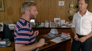 Een foto van presentator Tim. Hij staat links in beeld. Rechts zie je de toenmalige minister-president Mark Rutte. Tim is iets aan het vertellen. Achter hen zie je een bureau met stapels boeken erop. Daarachter zie je nog meer boeken staan. 