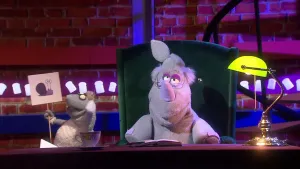 Een foto van de pop van Nico de Neushoorn op een groene stoel achter een bureau met een boek erop. Naast Nico is de pop Stella Schaap te zien. Stella Schaap heeft een bord vast met daarop een getekende slak met een slakkenhuis. 