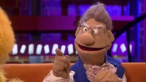 Een foto van de pop juffrouw Verbeten. Ze heeft een lichtbruine huid, grijs haar en ronde, streng kijkende ogen. Ze draagt een bril aan een koort dat naar beneden loopt naar haar nek. Ze wijst met haar rechterwijsvinger in de richting van de presentator.