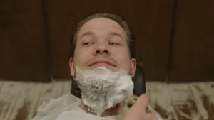 Een foto van het hoofd van Pascal die op een zwarte stoel naar achteren gebogen zit. Hij heeft wit scheerschuim op zijn kin en onder zijn mond zitten. De scheerkwast waarmee iemand het op zijn huid smeert is zichtbaar in een stukje van zijn of haar hand.