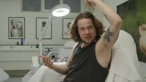 Een foto van Pascal die op een stoel met een witte handdoek erop zit. Pascal heeft zijn linkerarm boven zijn hoofd geheven. Zijn oksel is te zien en er zit geen haar onder zijn oksel. Achter Pascal staat een witte behandelstoel met een lamp erboven.