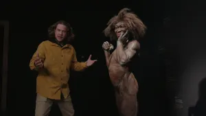 Een foto van Pascal die zijn handen met de handpalmen omhoog voor zijn lichaam houdt. Hij staat naast een echt lijkend beeld van een vrouwelijk oermens. Ze is naakt en heeft een bos bruin haar en een huid met zanderige vlekken. Ze krabt op haar wang.