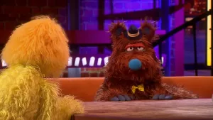 Een foto van de pop Brutus Bonk. Hij heeft een bruinharige vacht, een blauwe neus en zwarte nagels. Hij draagt een kleine zwarte hoed met gele randen tussen zijn oren. Ook draagt hij een gele strik. Links vooraan is de gele rug van de presentator te zien.