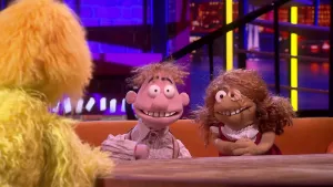 Een foto van de poppen Hans en Grietje aan een tafel. Ze hebben een grote grijns met ontblote tanden en grote ogen. Hans heeft opgetrokken wenkbrauwen. Hans draagt een blouse en bretels, Grietje een rood met witte jurk. Vooraan zit de presentator.