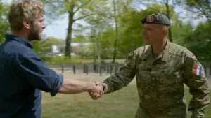 Een foto van presentator Tim. Hij schudt een militair de hand. Ze kijken elkaar aan. De militair heeft een groen uniform aan en een pet op. Op de achtergrond zie je bomen.