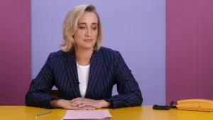 Een foto van Eva Jinek die achter een bureau zit. Voor haar liggen papieren en een pen. Naast haar staat een telefoon. Ze kijkt wat opzij en naar beneden.