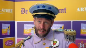 Een foto van Tim. Hij heeft een uniform aan en een pet op. Hij wordt geïnterviewd. Op de achtergrond zie je een bord met allemaal teksten erop. Bijvoorbeeld de tekst "De Grote Leider". 