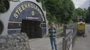 Een foto van presentator Janouk. Ze staat voor de ingang van een steenkolenmijn en praat naar de camera. Boven de ingang van de mijn staat de tekst "Steenkolenmijn. De allerlaatste in Nederland". Naast die ingang staan allemaal bomen.