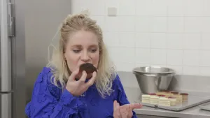 Een foto van presentator Janouk in een keuken. Ze neemt een hap van een gebakje. Op de achtergrond zie ongeveer tien kleine gebakjes op een aanrecht staan.