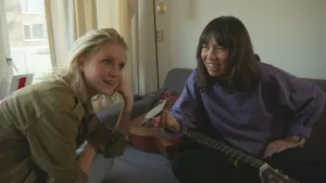 Een foto van presentator Janouk en Meau. Ze zitten in een woonkamer. Meau houdt een mobiele telefoon bij het hoofd van Janouk. Janouk luistert aandachtig. Er ligt een gitaar op de bank. 