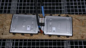 Een foto van een platte accu. Hij ligt op een houten plank. Die plank ligt op een ijzeren rooster. Aan de accu zijn twee dikke stroomdraden bevestigd. 