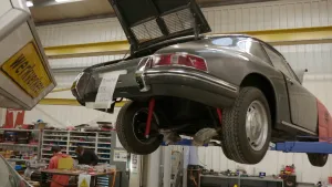 Een foto van een oude Porsche. Hij staat op de brug in een autowerkplaats. Je ziet de onderkant van de auto. Die is deels open gemaakt. Op de achtergrond zie je automonteurs.
