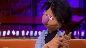 Een foto van de pop Pieter Pops die op een lichtbruine bank zit. Pieter heeft een zwarte mobiele telefoon in zijn handen. Pieter draagt een lichtblauwe blouse op een zwart t-shirt. Hij heeft zwart haar, een lichtbruine huid en een grote, lichtpaarse neus.