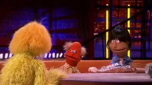 Een foto van drie poppen. De poppen aan de tafel zijn Hans en Kiki Gruismakers en de rug van de presentator. Voor Hans en Kiki liggen voorwerpen die ze bij een hoorspel gebruiken: een hamer, een wc-rol, kopjes en schoteltjes, een rieten mandje en folie. 