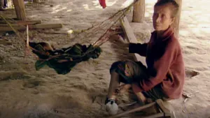 Een foto van een persoon die op een balk zit. De grond is van zand. Naast deze vrouw hangt een hangmat met daarin een baby. De vrouw krabt aan haar onderbeen. 