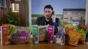Een foto van de presentator die achter een bureau zit. Hij is aan het bellen. Op het bureau ligt allemaal verpakt voedsel. Op een van die verpakkingen staat "veggie wraps".